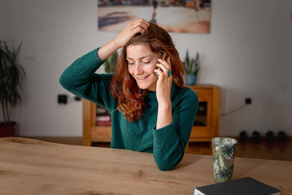 unge Frau telefoniert entspannt in ihrer Wohnung, trägt einen grünen Pullover und sitzt an einem Holztisch mit dekorativem Becher, natürliches Lächeln im wohnlichen Ambiente